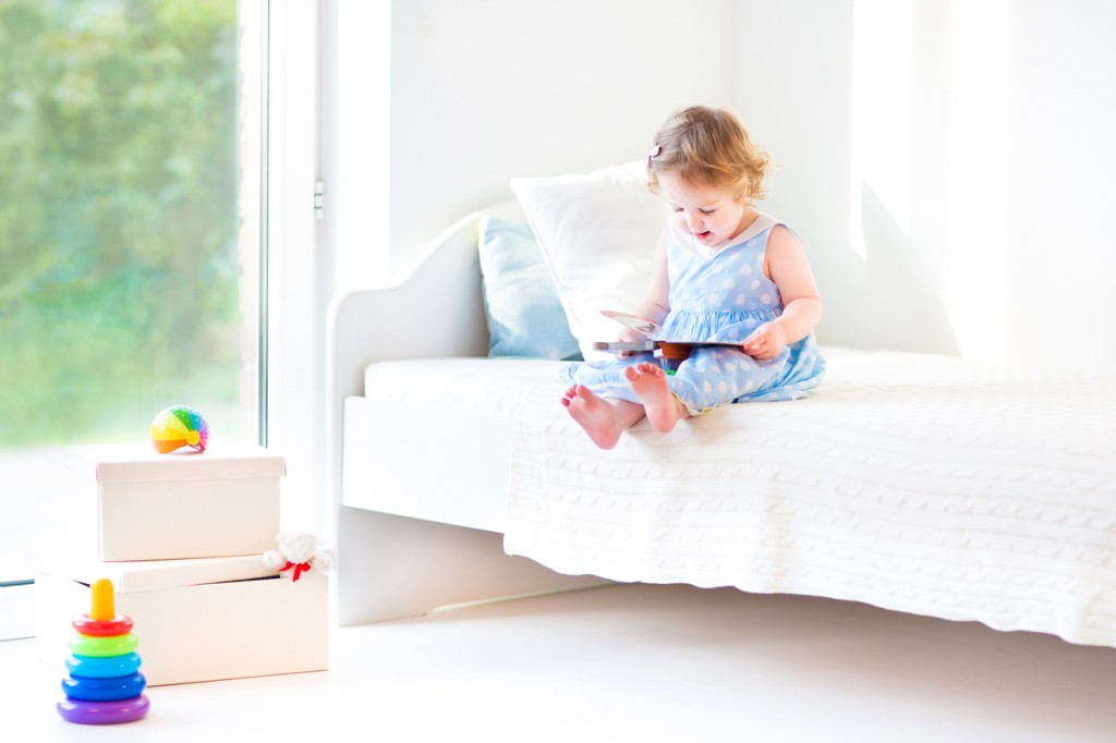 Adorable toddler girl reading book sitting on white bed